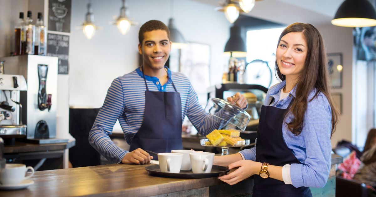 Two people in aprons at a café smiling at the camera. One is behind the counter lifting a lid off a tray of baked goods while the other is in front of the counter, holding a tray of coffee mugs.